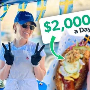 Amelia Eudailey, founder of Chef Hej Hej, smiling and flashing peace signs at her Swedish hot dog pop-up stand, with a fully loaded hot dog in the foreground.