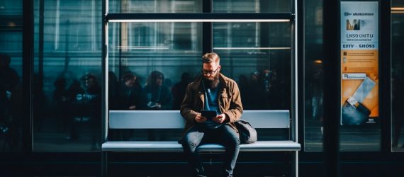 A man sitting at the bus stop taking surveys on his phone.